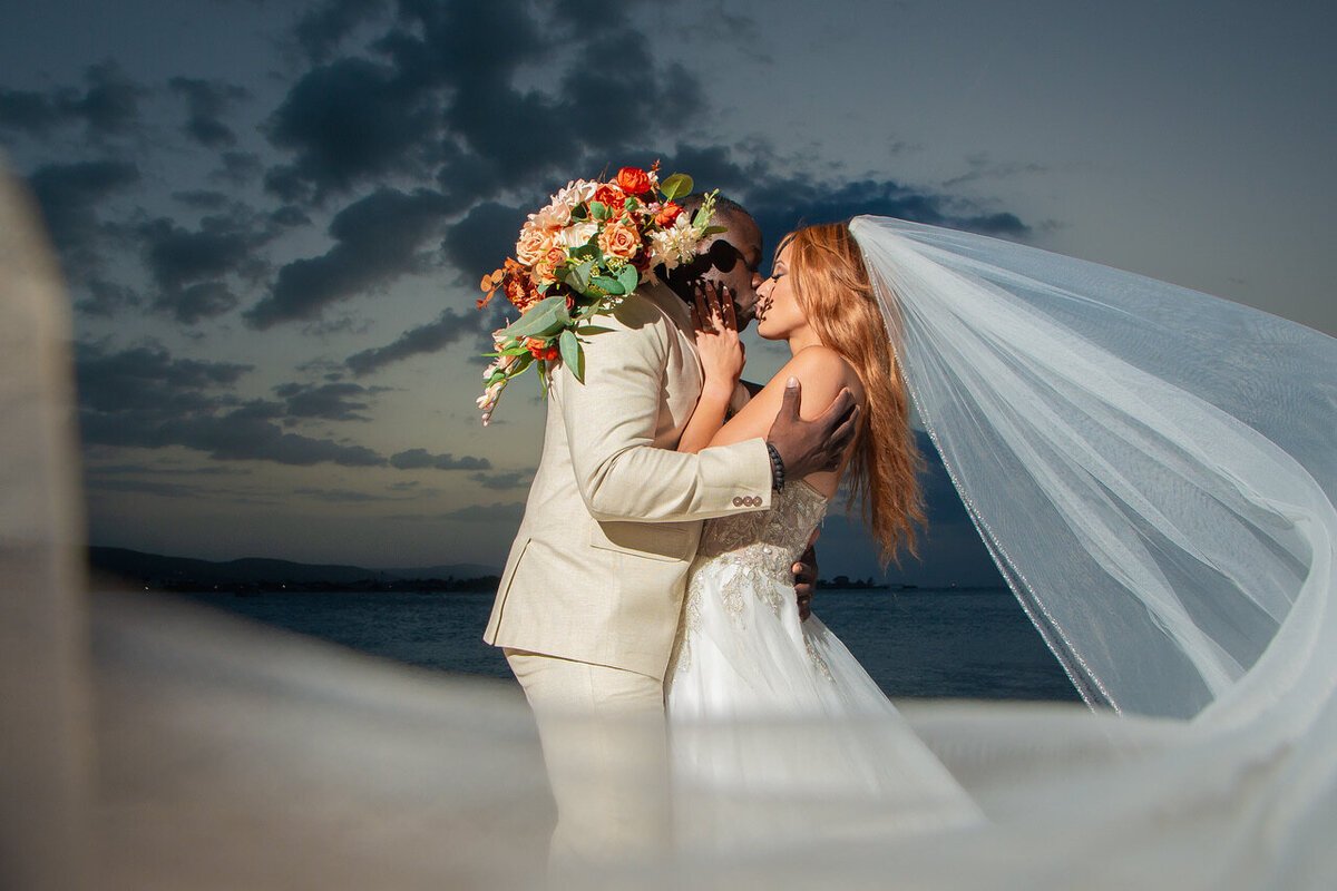 Couple embracing by the water at dusk, the bride's veil in the wind