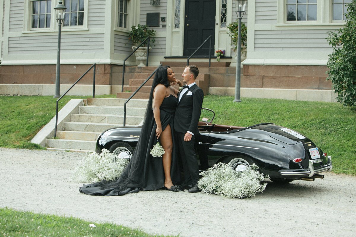 Couple in black wedding attire beside a vintage car