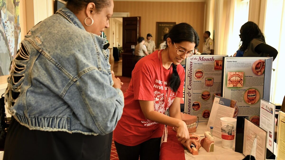 A student in a red Boston University shirt demonstrates a dental model to an attendee at a health booth